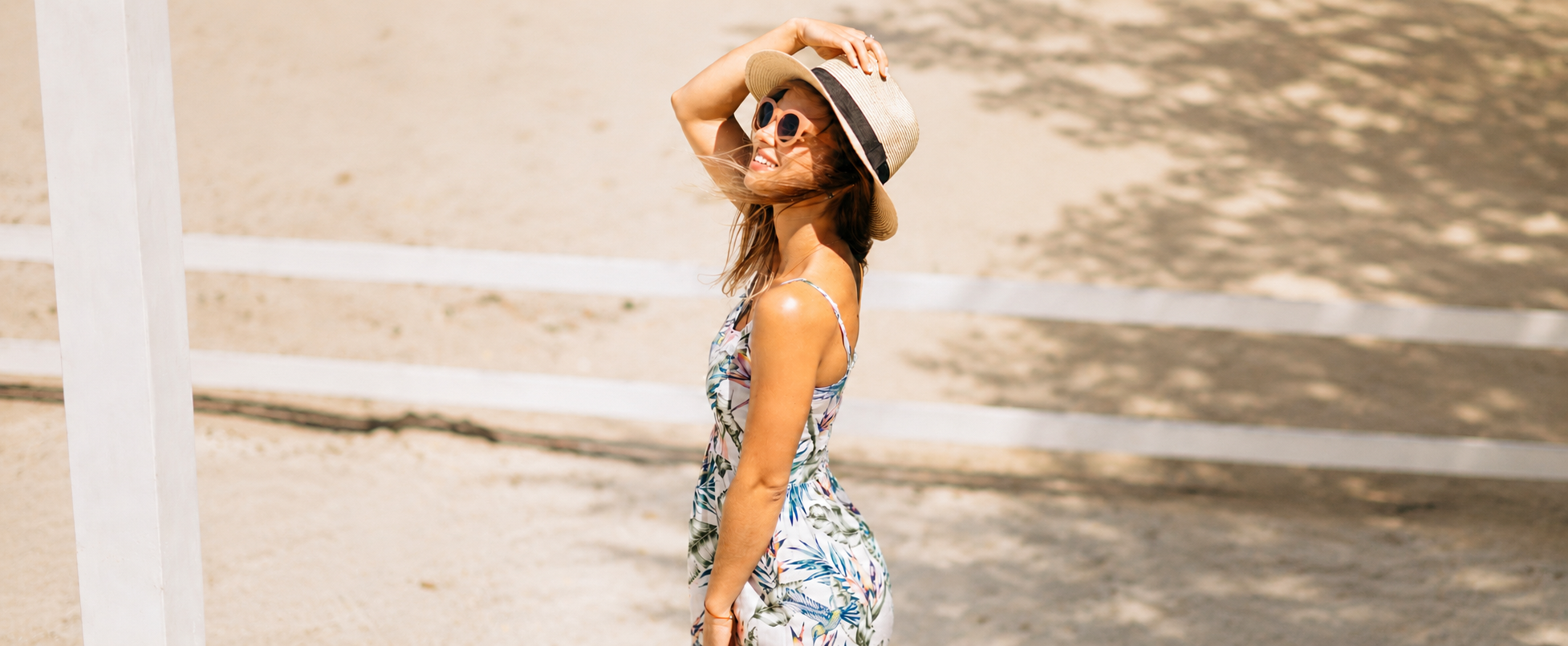 Woman in a floral dress and straw hat standing on a sandy beach.